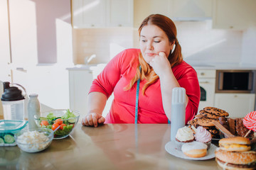 Fat young woman in kitchen sitting and eating food. She look at healthy meal on left side. Soft measure tape around neck. Sweet junk food on right side. Daylight in kitchen.