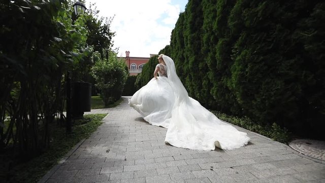 camera approaches bride in long wedding dress with lacy veil running between park trees and turning around slow motion