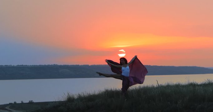 graceful young woman with long chiffon sheet preforms on green hill at calm river against red sunset light