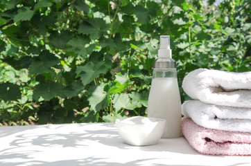 Laundry powder, softener and stack of clean towels on the white surface against green leaves.Natural light and leaves shadows