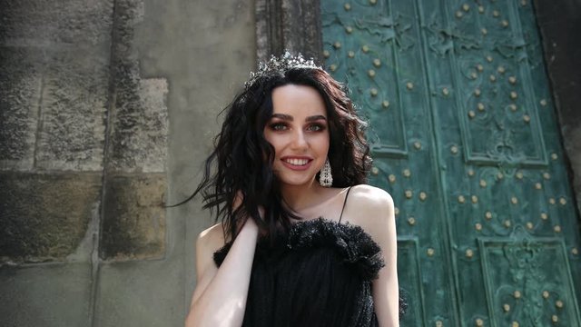thin spectacular girl, brunette, in long black dress, with bright make-up, crown on her head, posing, smiling, beckons to her, on the street near the old building, with high green doors, slow shooting