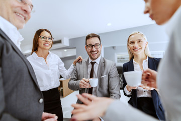 Group of smiling cheerful colleagues sitting and standing at workplace, chatting and drinking...