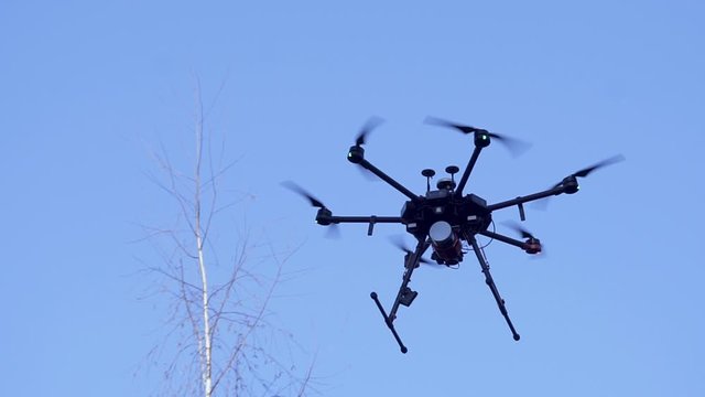 Black camera drone in flight with visible propelers movement and flashing light on blue sky background. Clip. Small quadcopter releasing the chassis against clear blue sky, slow motion.