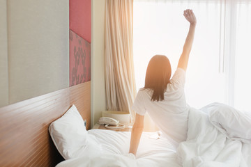 Woman stretching in bed after waking up, back view. Woman sitting near the big white window while stretching on bed after waking up with sunrise at morning, back view.