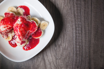 Closeup Vanilla ice cream poured with strawberry or raspberry jam with sliced banana slices on a white plate on a dark wooden table. You can do this at home on vacation