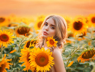 A young blonde woman stands in a field with blooming sunflowers and hugs a bouquet of flowers. Field mermaid at dawn. Background yellow field and fiery sunset. Photo with the addition of grain. - Powered by Adobe