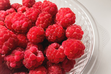 Close-up ripe juicy and delicious raspberry in a plastic transparent dish on a light background. Saturated healthy fresh food