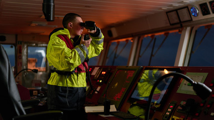 Navigator. pilot, captain as pat of ship crew performing daily duties with VHF radio, binoculars on board of modern ship with high quality navigation equipment on the bridge on sunrise.