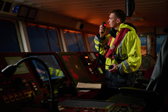Navigator. pilot, captain as pat of ship crew performing daily duties with VHF radio, binoculars on board of modern ship with high quality navigation equipment on the bridge on sunrise.