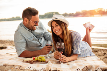 Beautiful young heterosexual couple enjoying picnic on a beach. Lying on a blanket, drinking red...