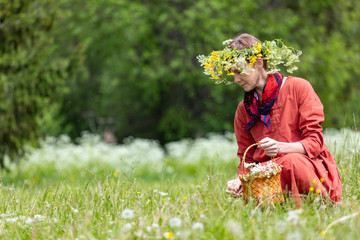 A girl in a national costume and a wreath on her head collects berries in a basket, on a green lawn in