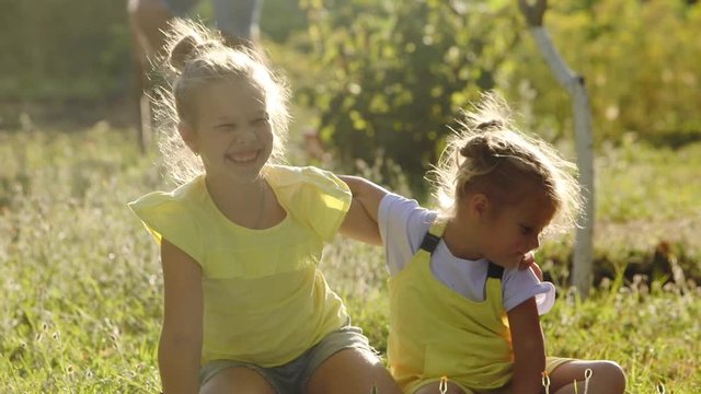friendly little blonde girls with space bubs sit on mat playing father walks with shovel in green garden on sunny day