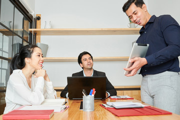 Young Asian businessman holding digital tablet and presenting the online presentation to his colleagues during business meeting at office