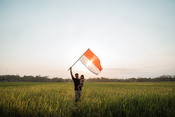 father carrying little girl pride flapping Indonesian flag with happiness in the rice field