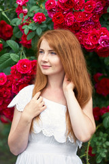 Portrait of a pretty redhead girl dressed in a white light dress on a background of blooming roses. Outdoor.