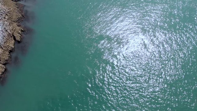 An aerial backward reveal footage of two islands rock, two kayakers and a rocky coastline with turquoise water under a majestic blue sky