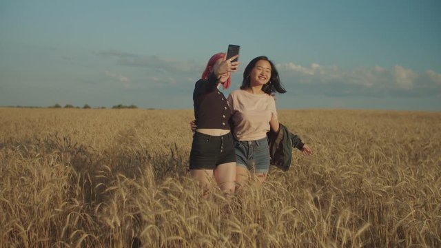 Excited positive multiethnic women taking selfie on smart phone in wheat field at sunset. Cheerful carefree females posing for selfie shot, making funny faces and fooling around on summer vacations.