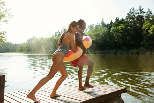 Happy friends having fun while laughting and jumping from the pier in river. Joyful male and female models in swimsuit at riverside in sunny day. Summertime, friendship, resort, weekend concept.