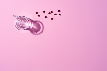 Glass cup with water and medicinal capsules on a pink surface. Top view.