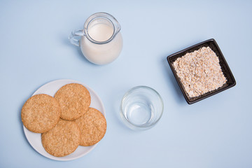 Leche de avena vegana sobre una mesa con un vaso de cristal, un bol con copos de avena y un plato de galletas.