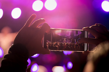 At a music festival, a man records his concert on his smartphone. There are no recognizable faces, only hands and stage lights.
