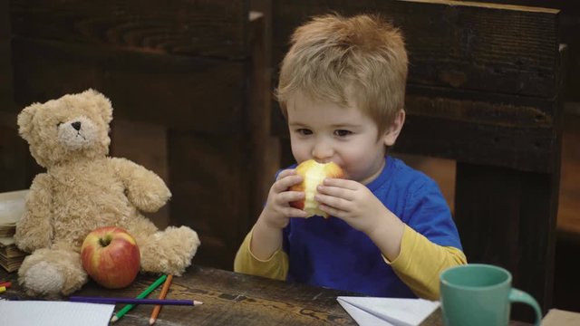 Cute blond child boy sitting in wooden chair and eating big red apple on wooden background. School break. Hungry kid biting apple in classroom. Small boy playing with paper plane and teddy bear.