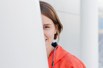 A short haired woman in coral windbreaker and colorful socks hiding behind a column near grey wall © Bohdan Melnyk