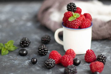 In a white, small mug are raspberry berries, mint leaves. This is a vitamin mug. She is standing against a dark background. Nearby are blackberries and flowers.