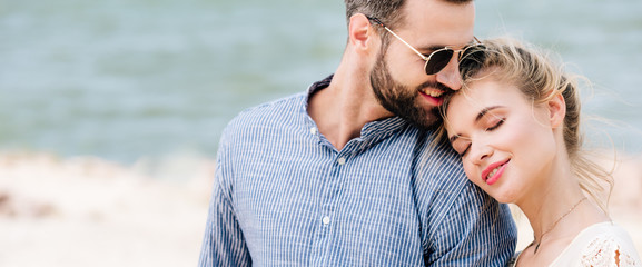 Fototapeta premium blonde girl with closed eyes leaning on boyfriend shoulder while man smiling at beach, panoramic shot