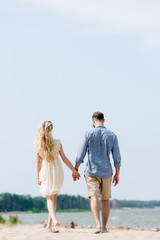back view of adult couple walking along beach and holding hands at sunny day