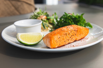 Grilled salmon steak on the side with arugula and salad. Served with a sauce and lime on a white plate in the restaurant.