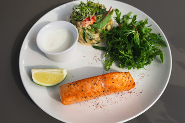 Grilled salmon steak on the side with arugula and salad. Served with a sauce and lime on a white plate. White sauce.