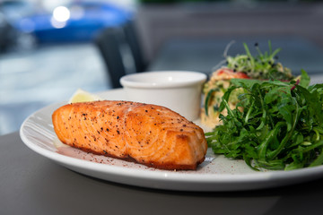 Grilled salmon steak on the side with arugula and salad. Served with a sauce on a white plate in the restaurant. Closeup with bokeh.