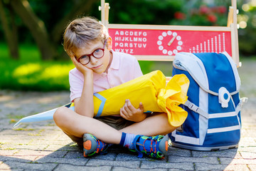 Frustrated sad little kid boy with glasses sitting by desk and backpack or satchel. Schoolkid with traditional German school bag called Schultuete on his first day to school. Hello school in German.