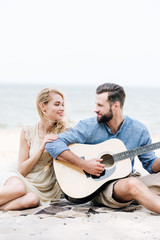 smiling beautiful young barefoot woman sitting on blanket and looking at boyfriend with acoustic guitar at beach near sea