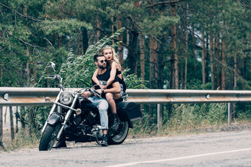 young couple of bikers hugging while sitting on black motorcycle on road near green forest