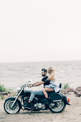 Fototapeta na wymiar side view of young couple of bikers riding black motorcycle at sandy beach near river
