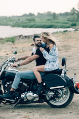 Fototapeta na wymiar smiling young couple of bikers hugging on black motorcycle at sandy beach