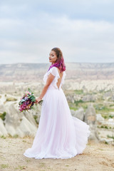 Fototapeta na wymiar Woman with a beautiful bouquet of flowers in her hands stands on the mountain in the rays of the dawn sunset. Beautiful white long dress on the girl body. Perfect bride with pink hair