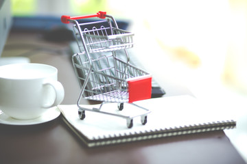 Online shopping concept.Shopping cart on desk with notebook and white cup of coffee