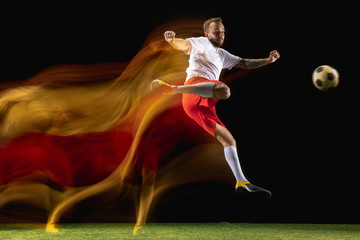 Concentrated. Young caucasian male football or soccer player in sportwear and boots kicking ball for the goal in mixed light on dark background. Concept of healthy lifestyle, professional sport, hobby