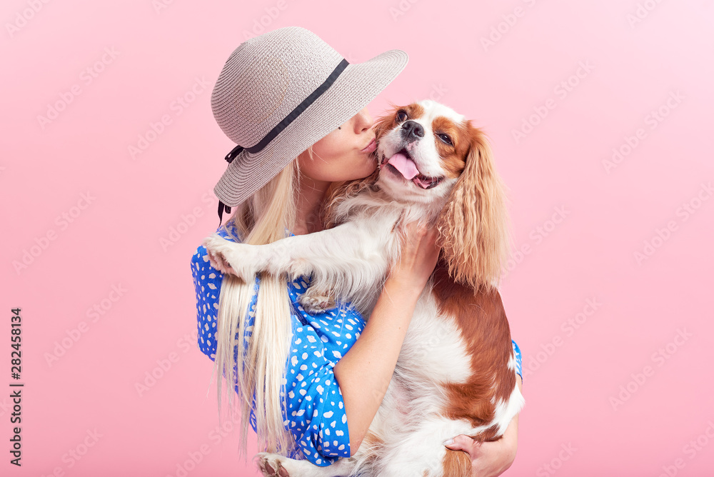 Wall mural Portrait of smiling young blond woman in summer hat embracing king Charles spaniel dog. owner and pet relations concept. Veterinary health. Isolated front view on pink background.