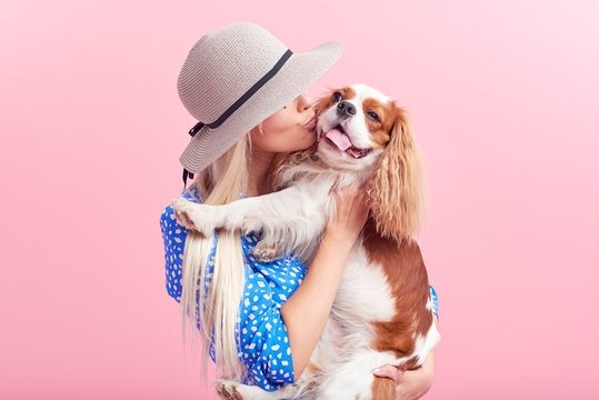 Portrait of smiling young blond woman in summer hat embracing king Charles spaniel dog. owner and pet relations concept. Veterinary health. Isolated front view on pink background.