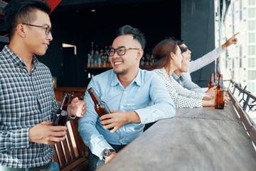 Two Asian friends drinking beer and talking to each other during their leisure time in the bar with other people in the background