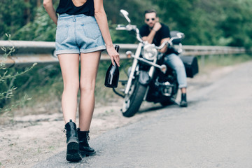 cropped view of young woman walking on road with bottle of alcohol near boyfriend on black motorcycle