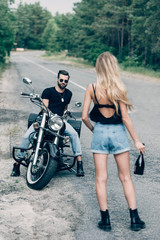 Fototapeta na wymiar back view of girl walking on road with bottle of alcohol near handsome boyfriend in sunglasses on black motorcycle