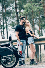Fototapeta na wymiar young couple of bikers embracing near black motorcycle on road near green forest
