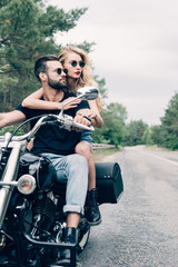 Fototapeta na wymiar young couple of bikers hugging and looking away on black motorcycle on road near green forest