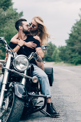 young couple of bikers closely looking at each other on black motorcycle on road near green forest