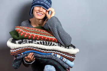 Young woman hold pile of sweaters wearing knitted hat and warm wool sweater